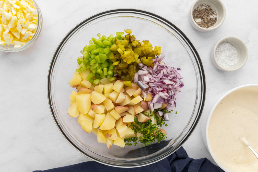 Chopped onions, celery, pickles, chives and boiled potatoes layered into a mixing bowl to make potato salad.