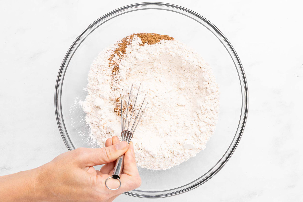 A hand stirring peach muffin dry ingredients in a bowl: flour, cinnamon, baking powder, and baking soda.
