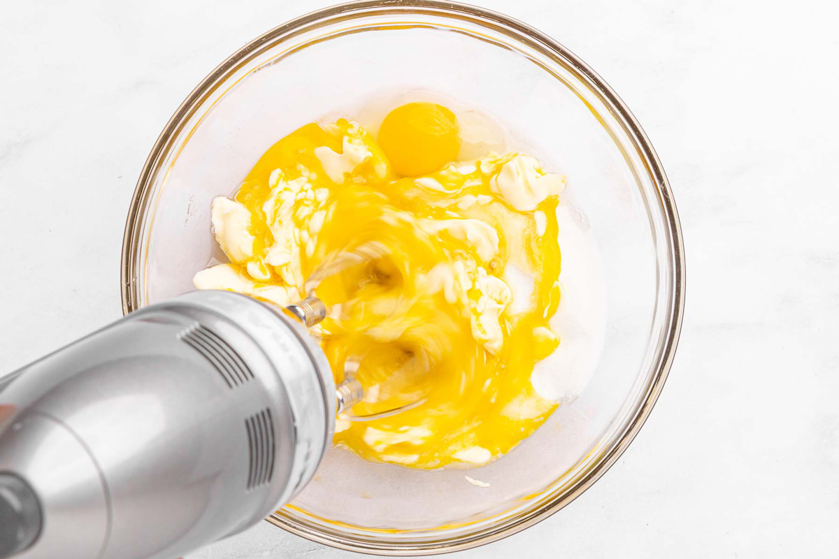 Hand mixer whisking peach muffin wet ingredients: eggs, vanilla, sugar, yogurt, and buttermilk.