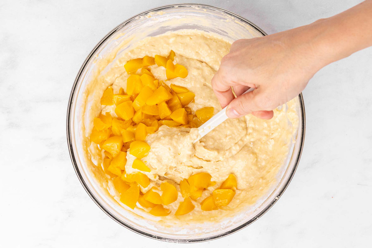 A hand holding a spatula folding peaches into peach muffin batter.