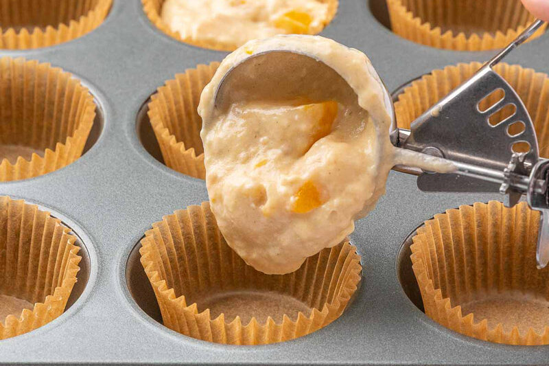 A cookie scoop full of peach muffin batter filling muffin cups.