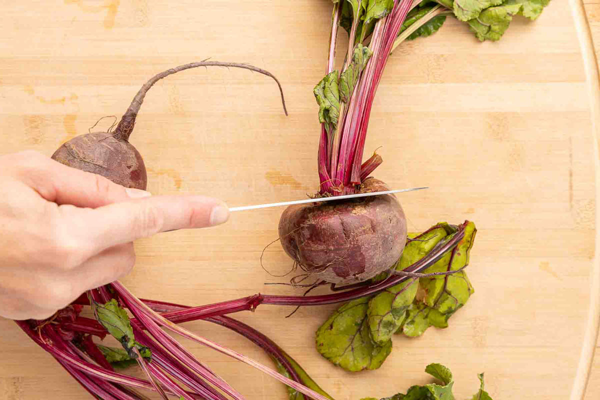 A hand using a knife to separate beets from their stems.