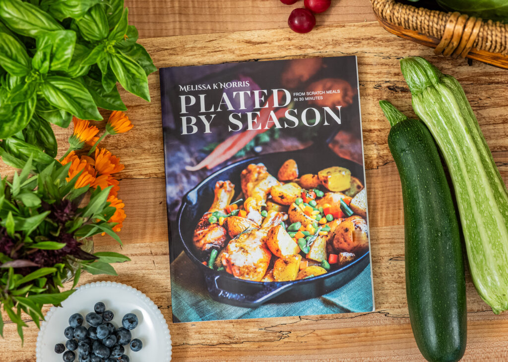 Plated By Season from-scratch 30 minute meals magazine on butcherblock countertop with zucchini, fresh basil, blueberries, and cabbage surrounding it.
