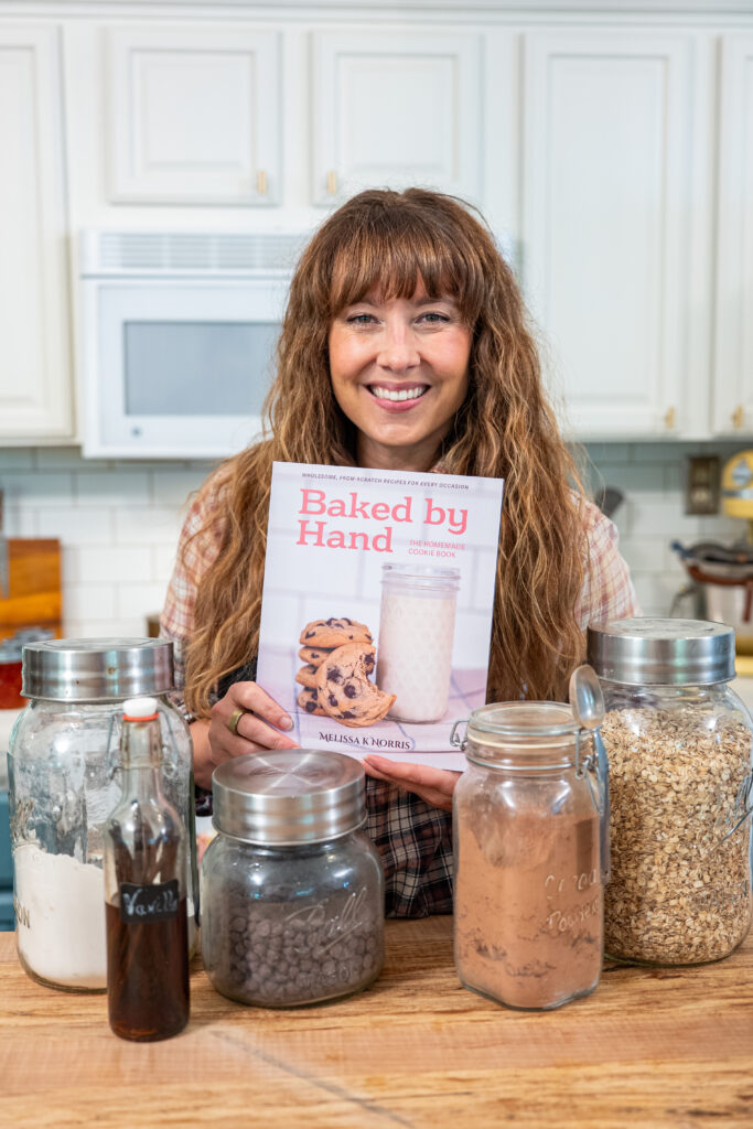 A woman holding a copy of Baked by Hand: homemade cookie book with jars of flour, vanilla extract, chocolate chips and cocoa powder in a kitchen