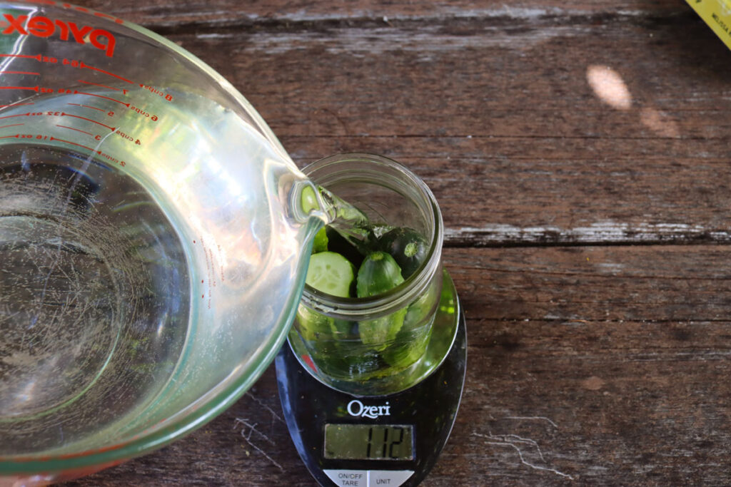 Pouring water into a jar of pickles sitting on a kitchen scale.