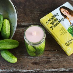A jar of fermented pickles sitting in between freshly washed cucumbers and Melissa's book 