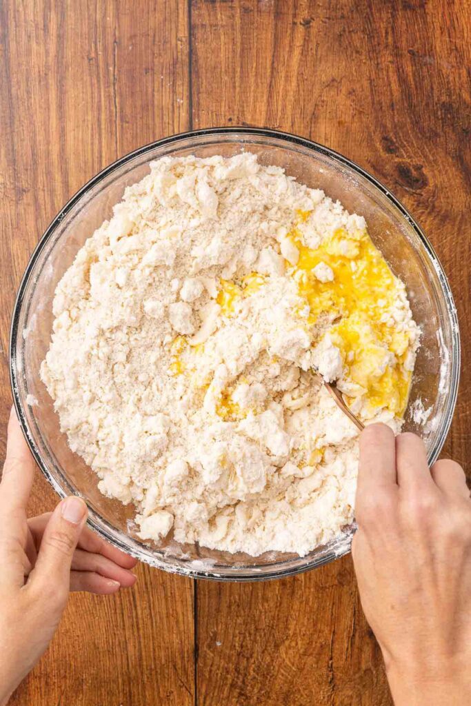 A hand using a fork to blend together pie crust ingredients in a glass bowl.