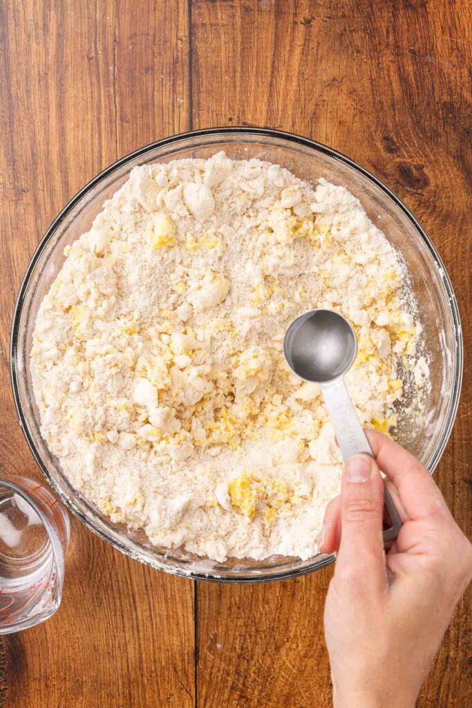 A hand adding a tablespoon of water to pie crust ingredients in a glass bowl.