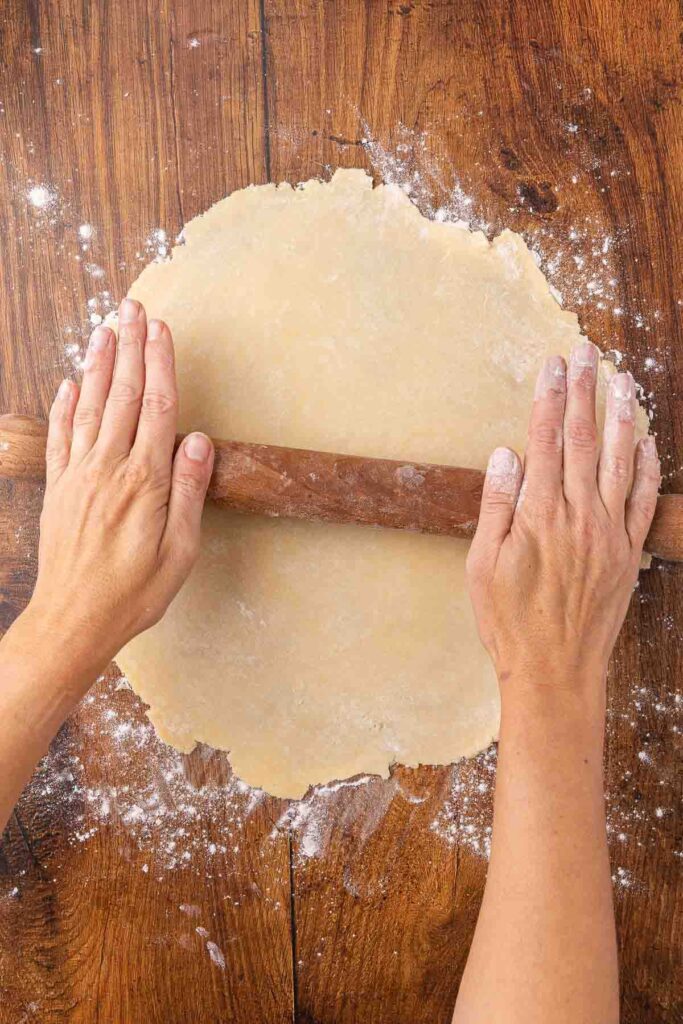 Hands using a rolling pin to roll out pie crust onto a floured surface.