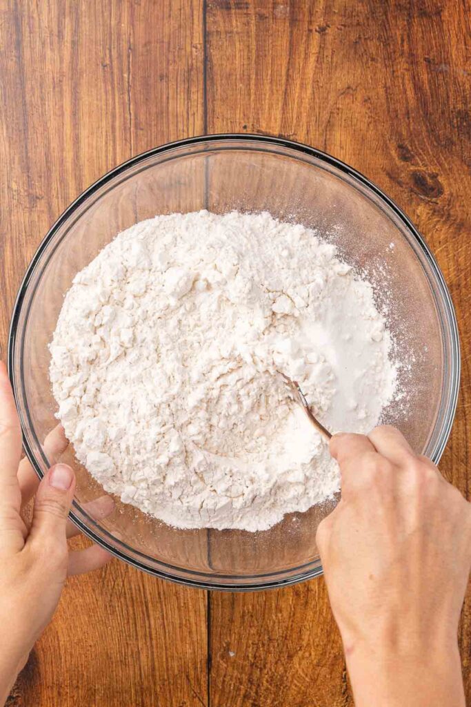 A hand using a fork to mix together flour, salt, and sugar in a glass bowl.
