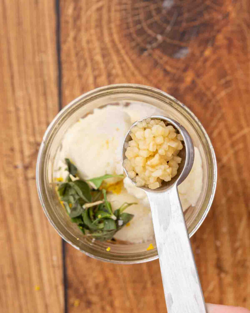 A spoon adding garlic to oil, herbs and cheese in a glass Mason jar.