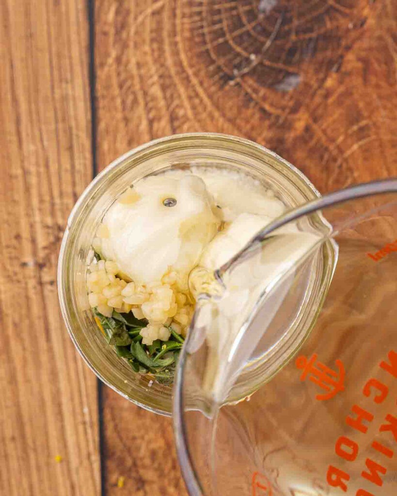 Oil pouring into a jar filled with mozzarella balls, herbs, lemon zest, lemon juice, and garlic.