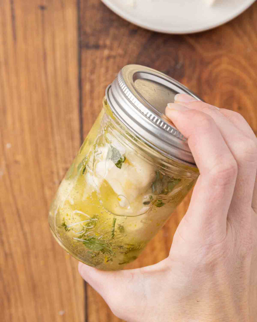 A hand shaking a sealed jar of mozzarella balls to combine ingredients.