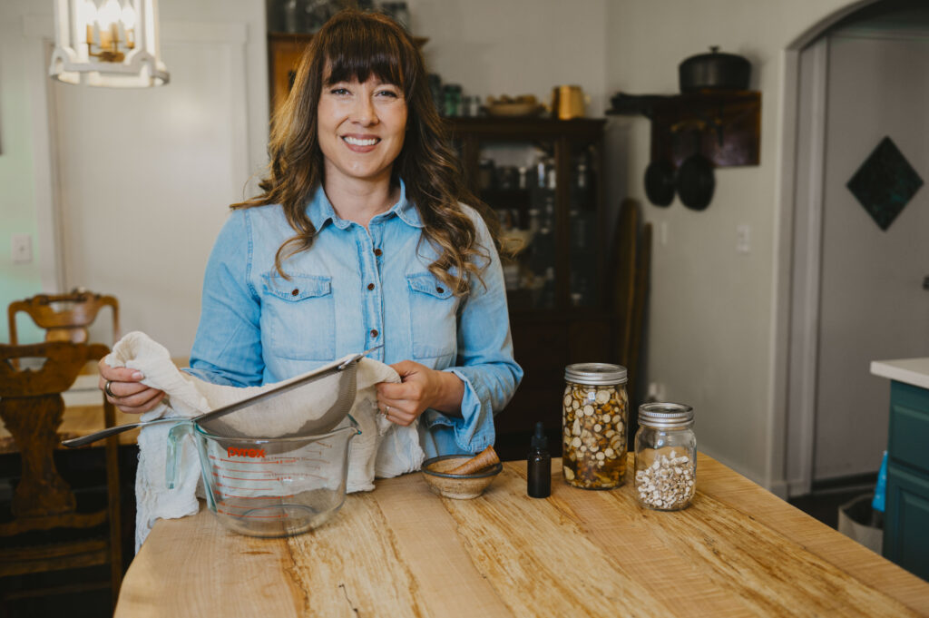 A woman standing at her kitchen island with jars of dried herbs, an amber tincture bottle and a pottery mortar and pestal straining an herbal tincture into a bowl.