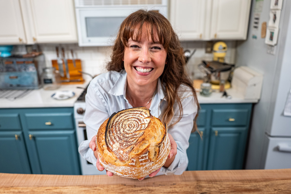 A woman holding up a sourdough boule loaf.