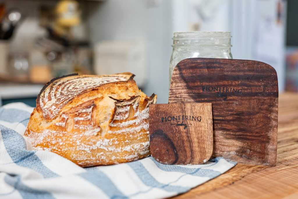 artisan sourdough loaf with a wooden bench knife and dough scraper and jar of sourdough starter on kitchen counter