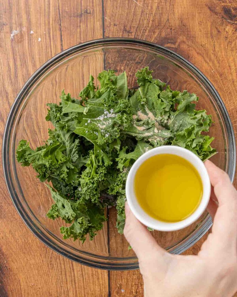 A hand pouring olive oil into a bowl of kale pieces.
