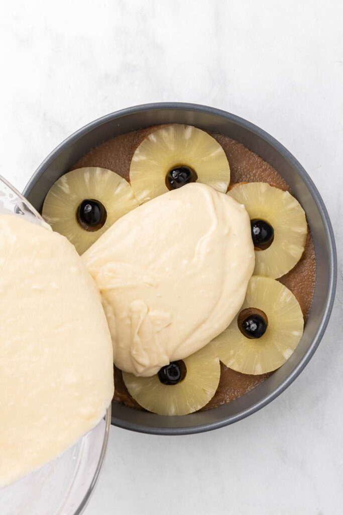 Pouring cake batter over cake pan prepared with brown sugar topping, slices of pineapple and cherries.