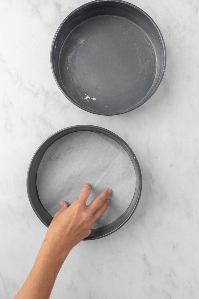 A hand placing parchment paper into the bottom of a nine inch round cake pan.