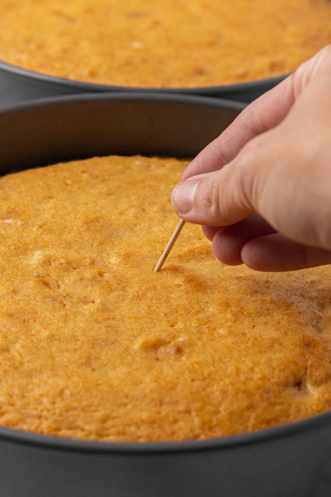 A hand piercing tangerine cake with a toothpick to check for doneness.