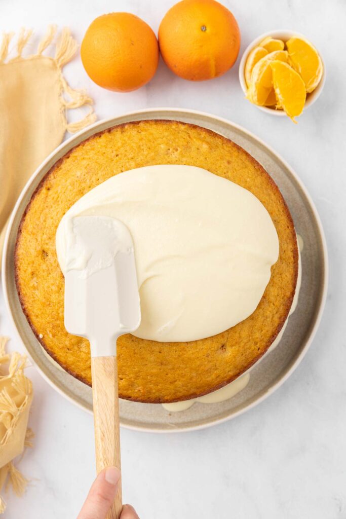 A spatula spreading frosting over the base layer of a tangerine cake.