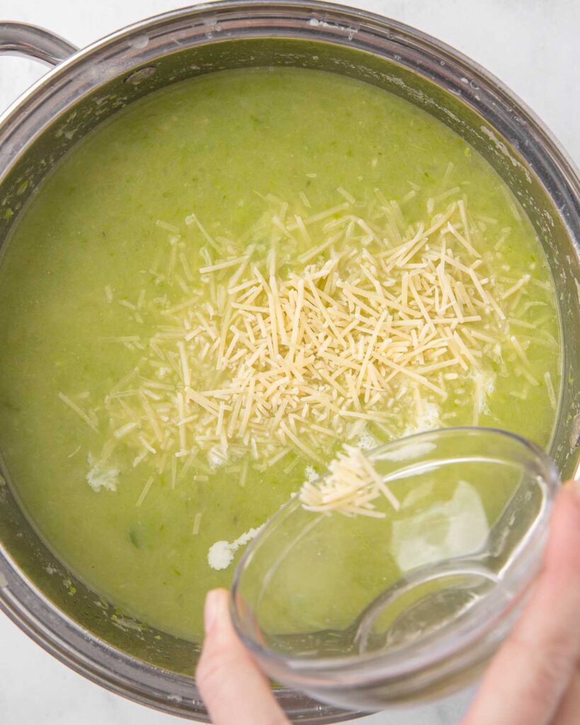Adding Parmesan cheese to pureed asparagus soup in a saucepan.