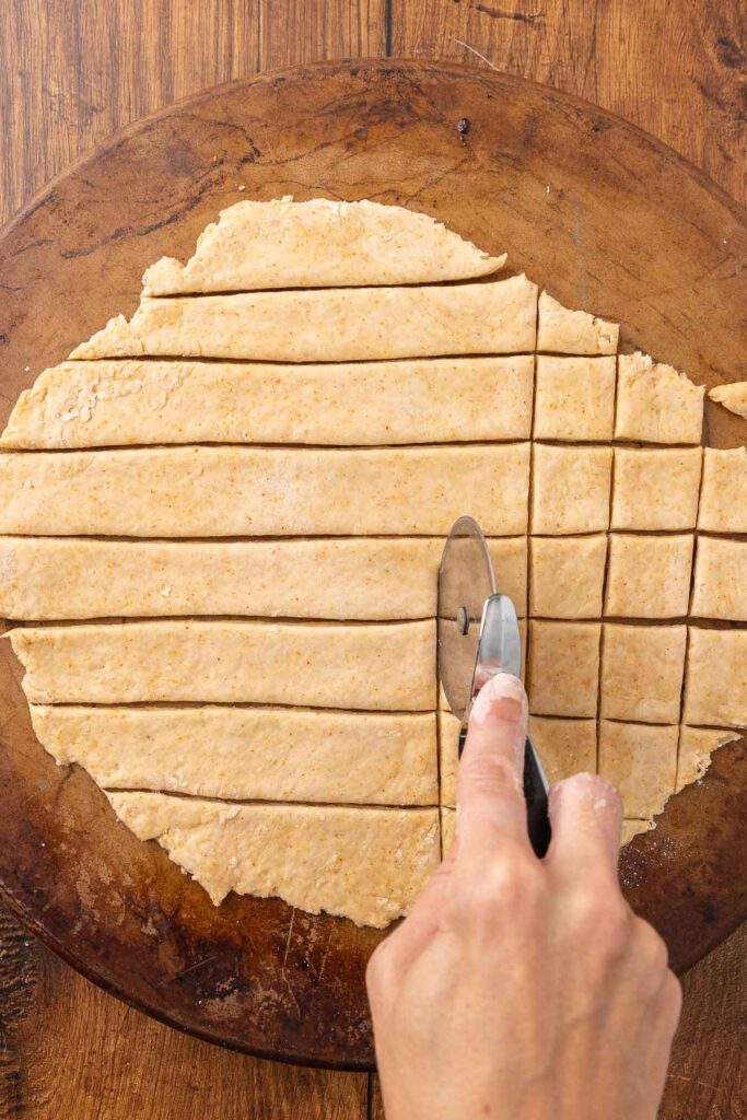Hands using a pizza cutter to cut cracker dough into pieces.