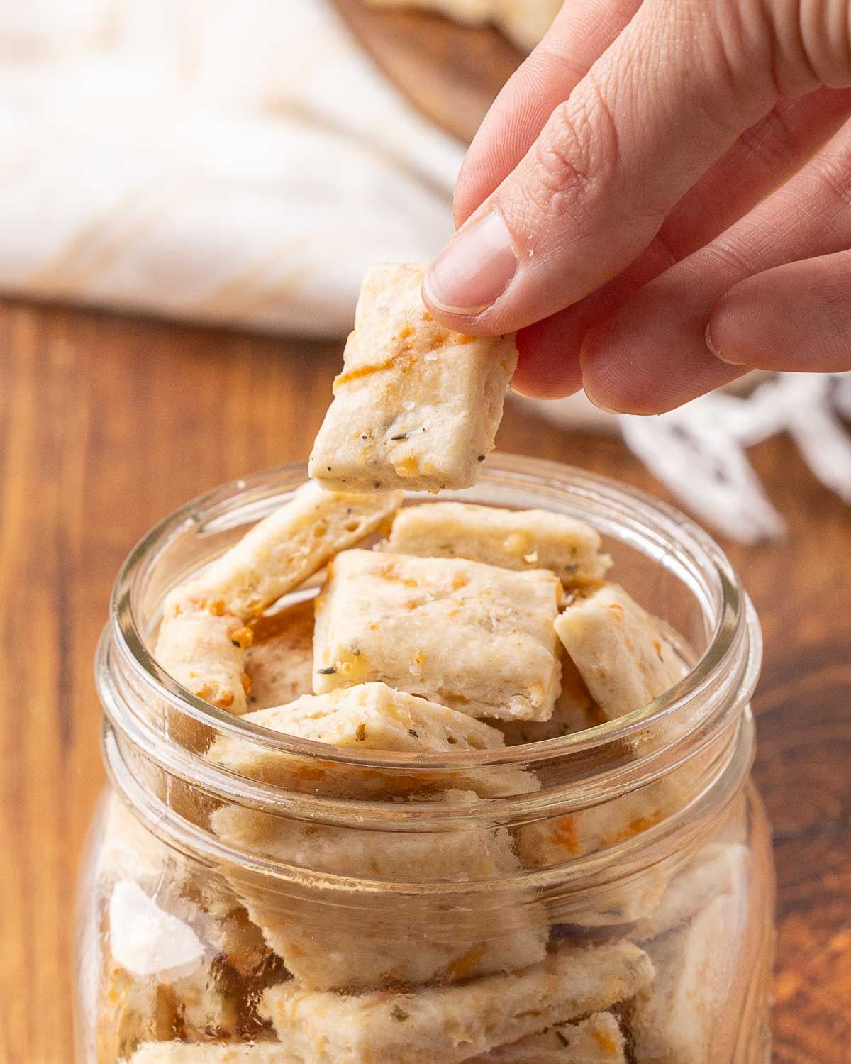 A hand pulling a sour cream and onion cracker from a glass jar.