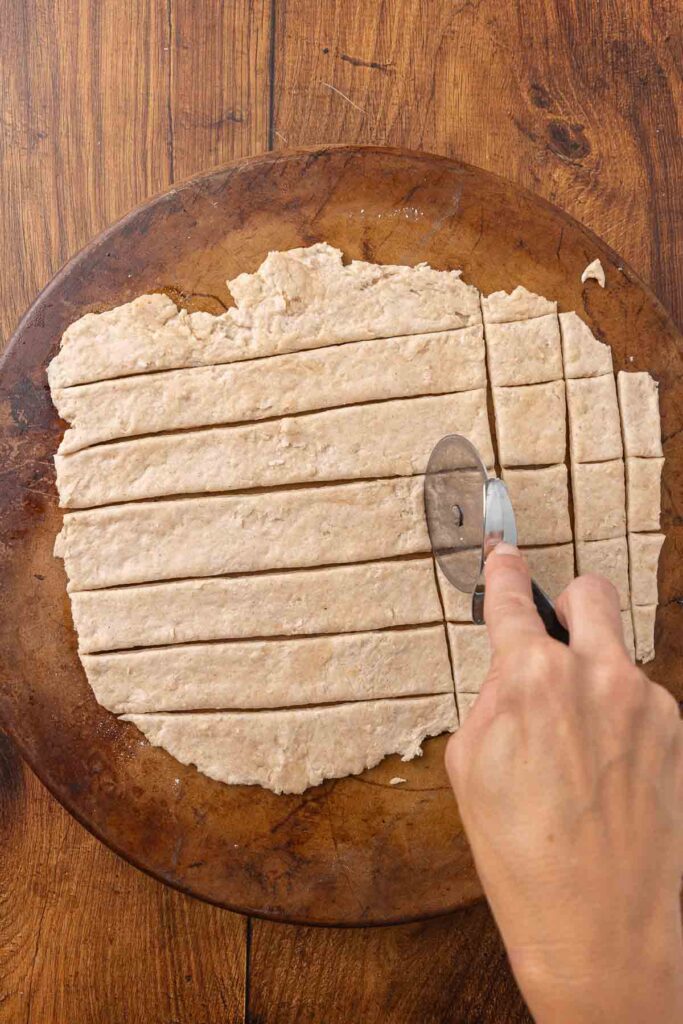 Hands using a pizza cutter to cut cracker dough into pieces.