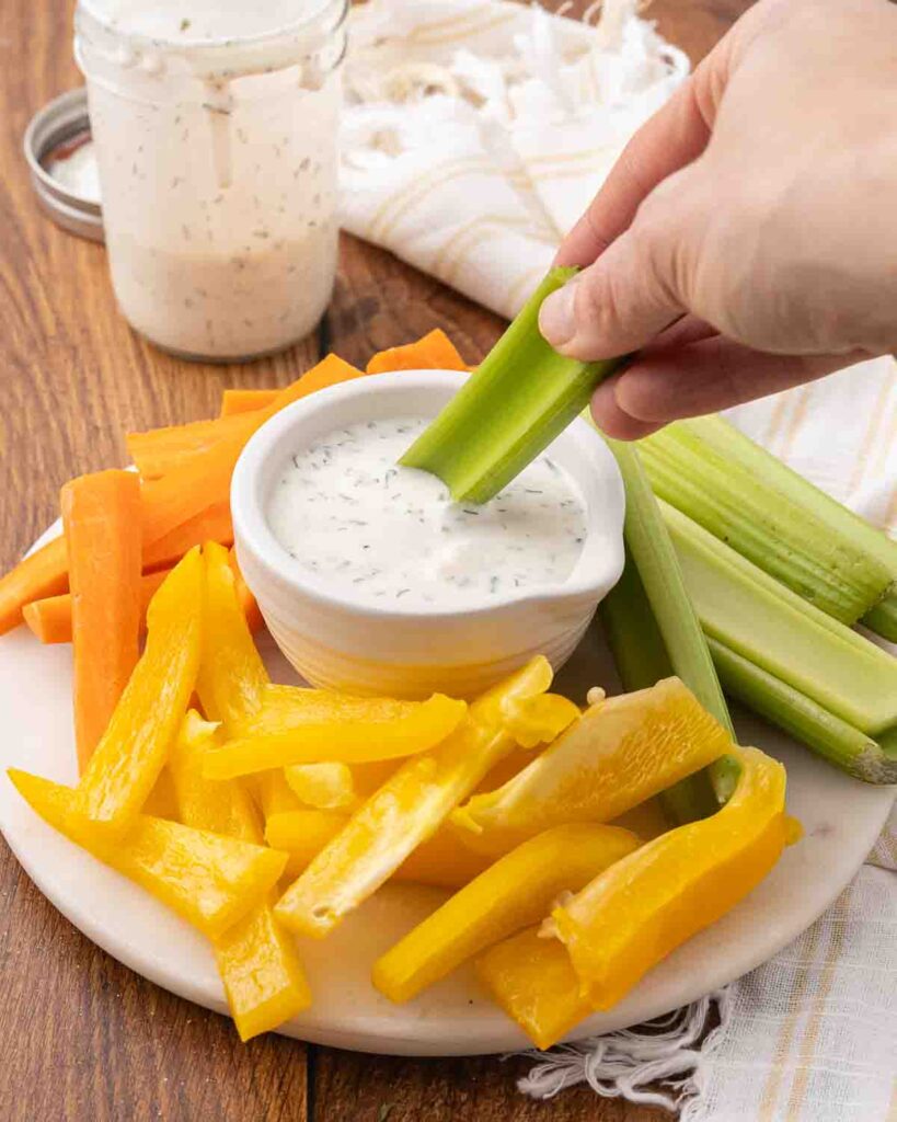Assorted sliced veggies on a platter with a hand dipping a celery stick into a bowl of homemade ranch dressing.