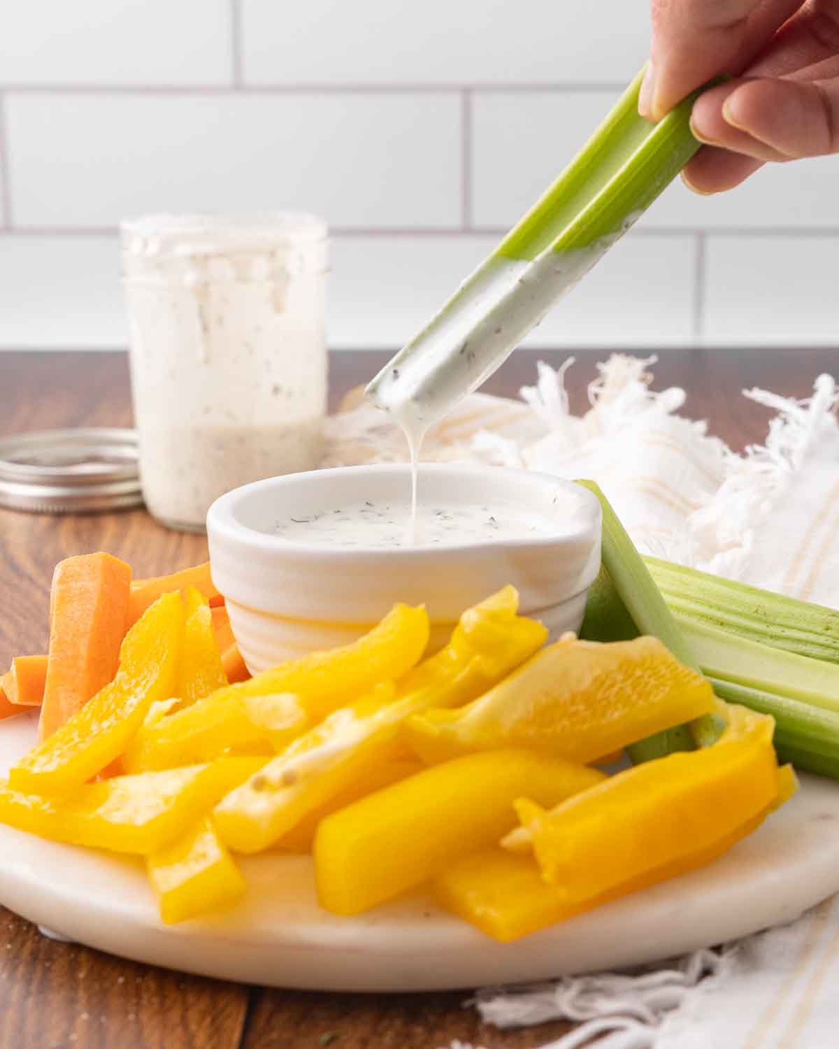 Assorted sliced veggies on a platter with a hand dipping a celery stick into a bowl of homemade ranch dressing.