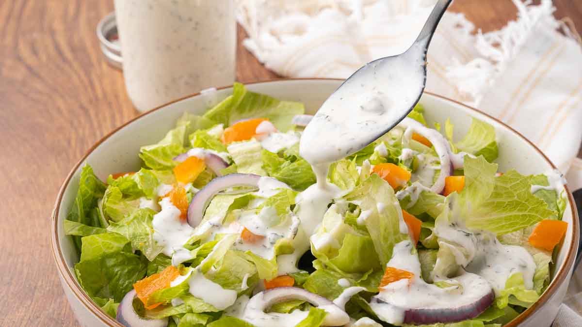 A bowl of green salad with a spoon drizzling homemade buttermilk ranch over the top.