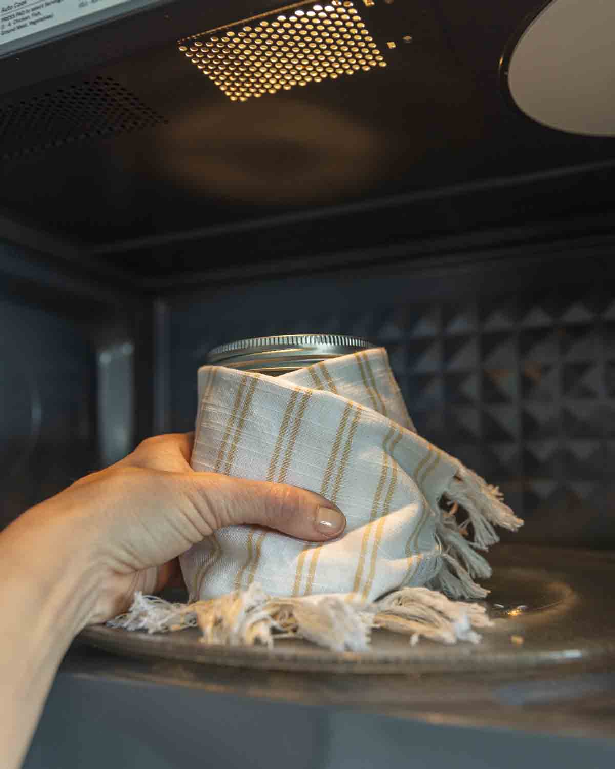 A hand placing a towel wrapped jar filled with milk and yogurt cultures into a microwave to maintain culturing temperature.