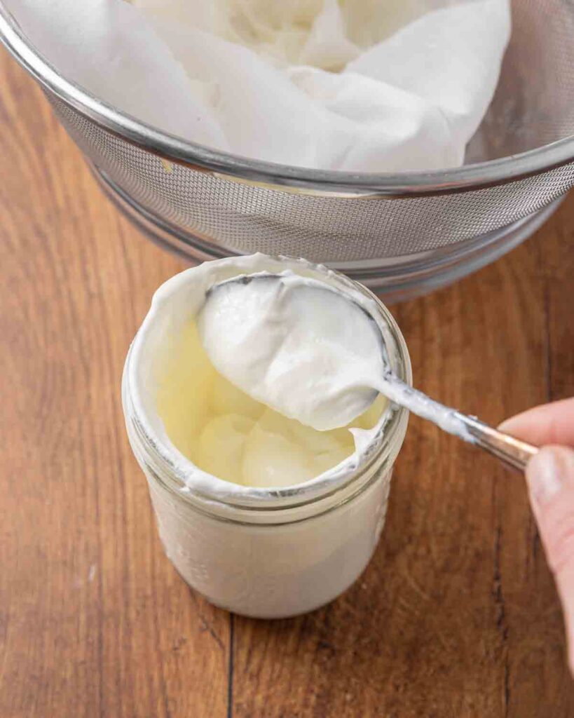A spoon transferring finished homemade Greek yogurt from the cheesecloth into a clean jar.