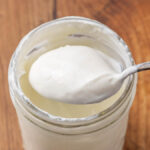 A jar of homemade Greek yogurt with a spoon lifting a bite to show texture.