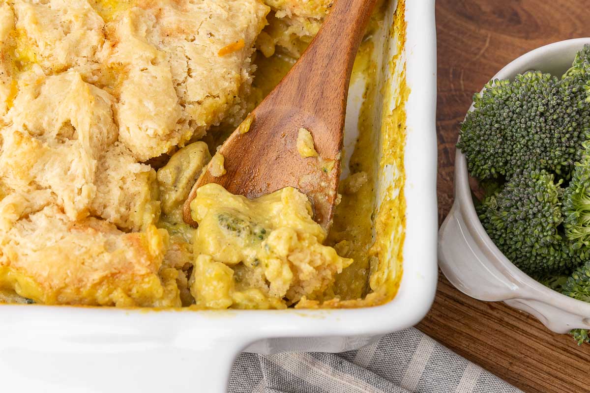 A bird's eye view of a chicken and broccoli casserole with a wooden spoon dishing out a serving.
