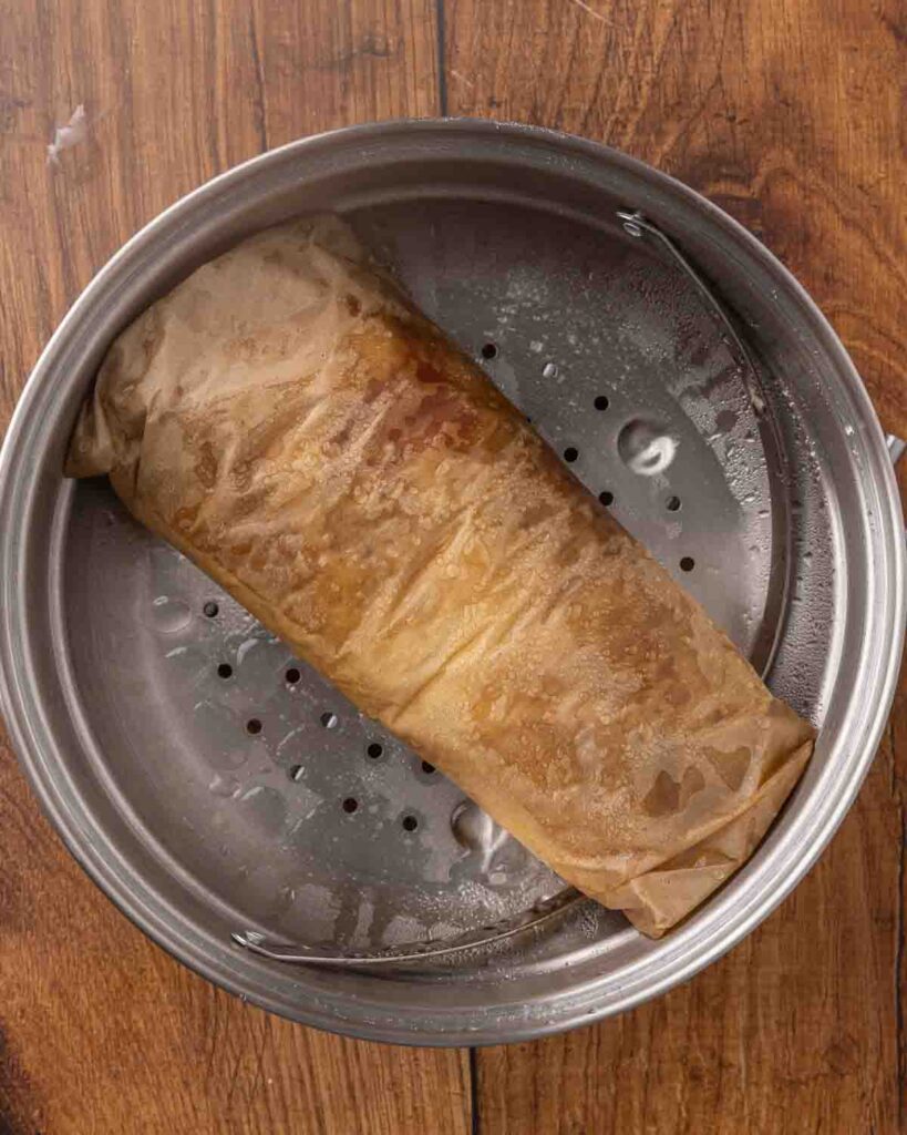 A jam roly poly wrapped in parchment paper, and steaming in a pan.
