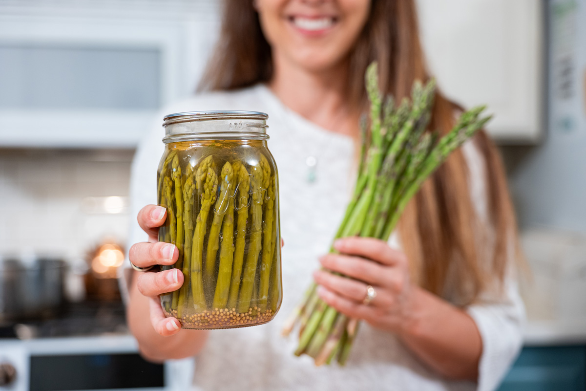 A woman holding up a jar of pickled asparagus and in the other hand fresh asparagus.