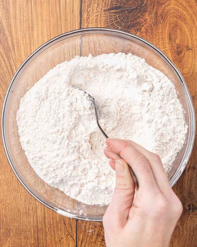 A hand holding a spoon stirring together a flour mixture in a glass bowl.