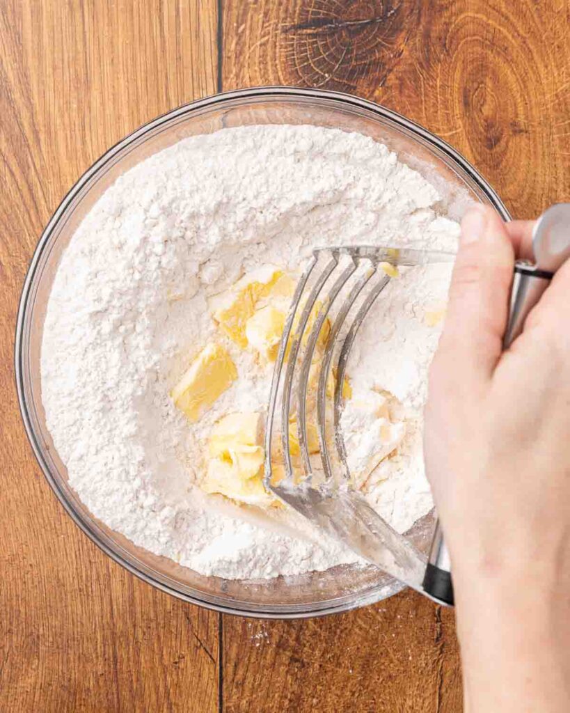 A hand holding a pastry cutter, cutting butter into flour mixture.