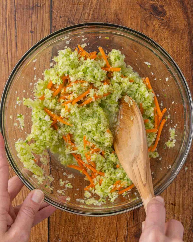 A wooden spoon mixing salt, shredded carrots, and shredded cabbage together in a bowl.