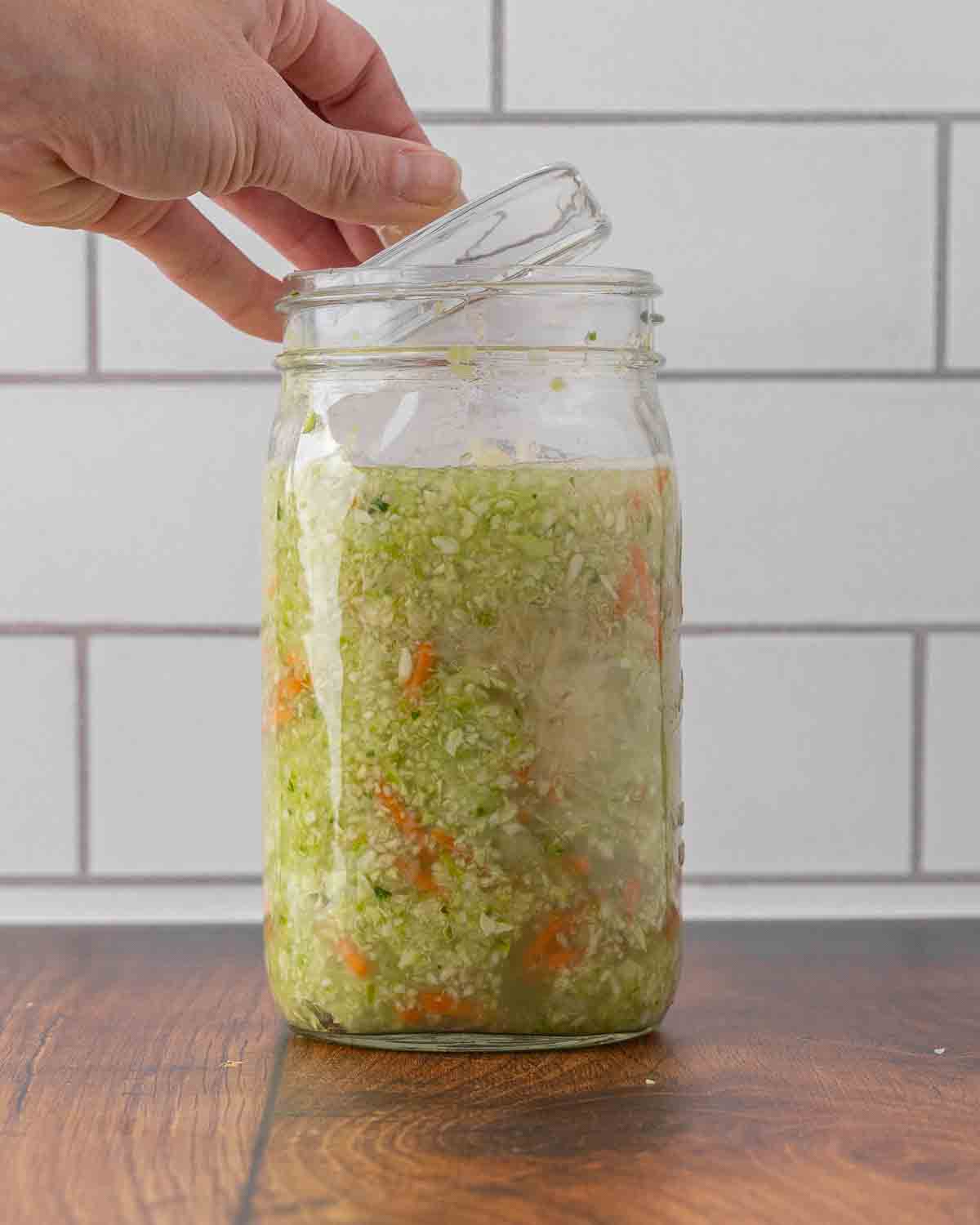 A hand placing a fermentation weight on top of a jar full of cabbage and brine ready to ferment.