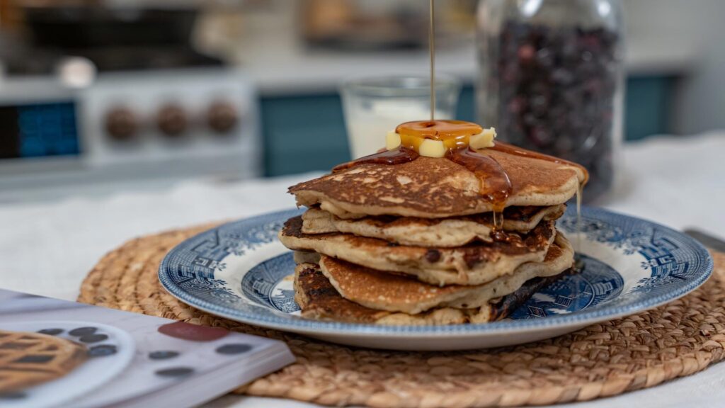 A stack of sourdough pancakes on a blue plate with maple syrup being poured over the top and a jar of freeze dried blueberries in the background.