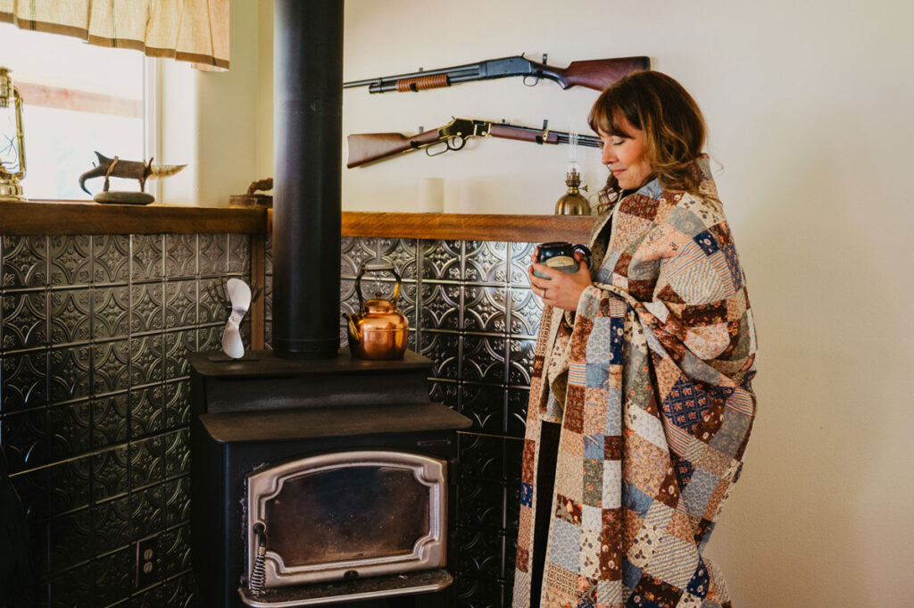 A woman standing in front of a wood stove, wrapped in a quilt, and holding a mug.