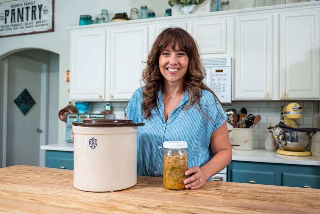 A woman standing by a fermenting crock and a jar of sauerkraut.